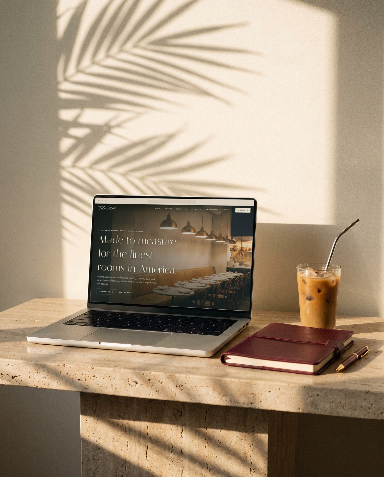 A laptop on a travertine desk in late sun showing the new Tailor Booth home page, an iced coffee and a burgundy notebook beside it.