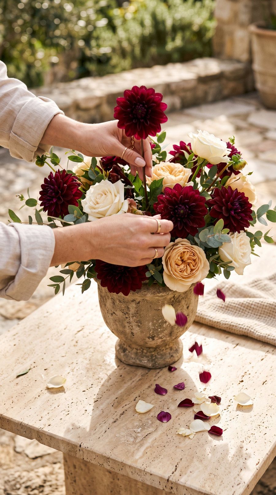 Hands arranging burgundy dahlias and cream roses in a stone vase on a sunlit table.