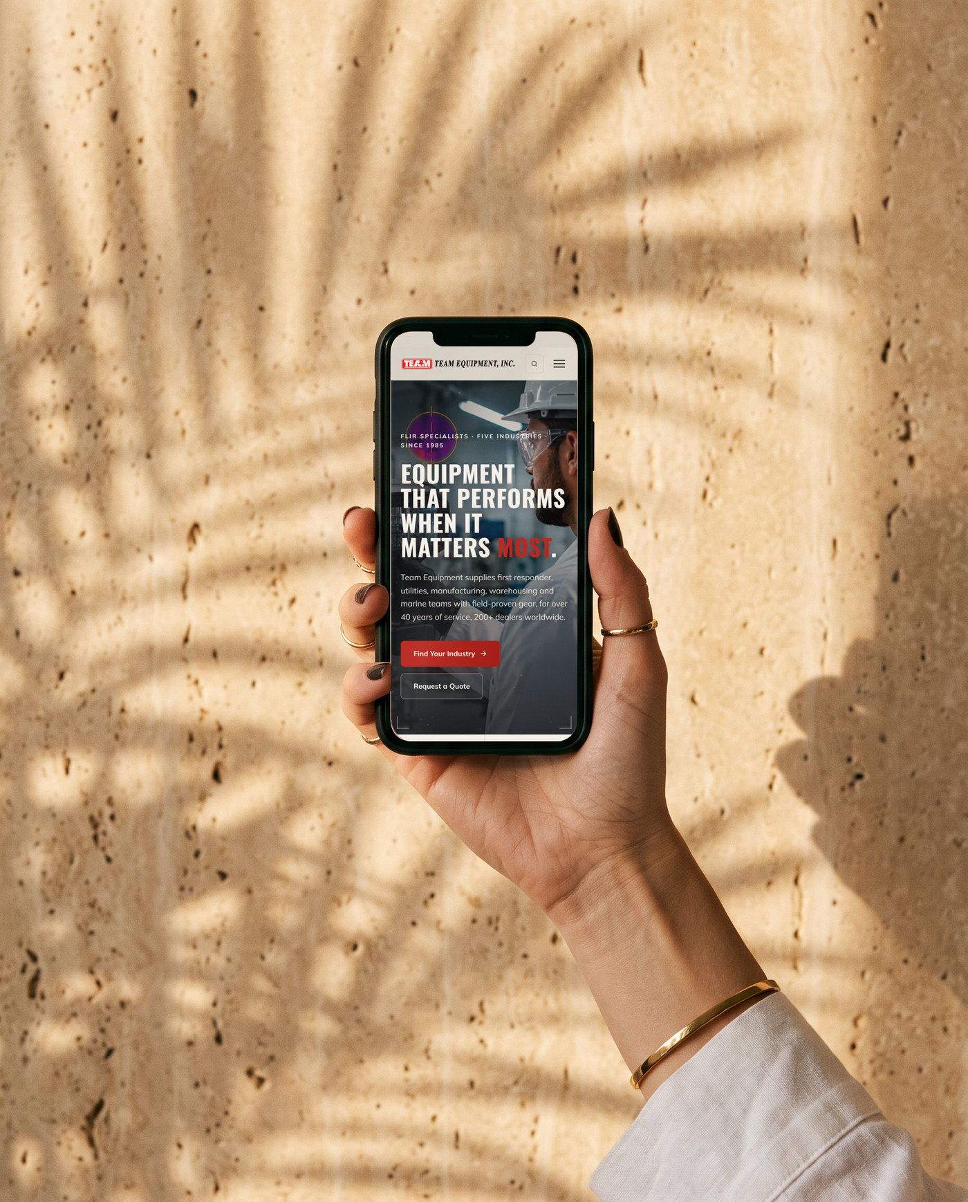 A hand holding a phone against a travertine wall, showing the new Team Equipment mobile home page.